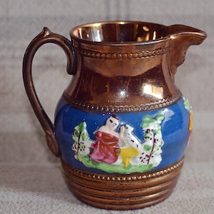 May include: A small, antique pitcher with a brown exterior and a blue band featuring a pastoral scene. The interior has a gold finish. The handle and spout are brown. The pitcher is likely made of ceramic.
