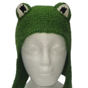May include: A green knitted hat featuring two protruding frog eyes on top. The eyes are white with black pupils. The hat has ear flaps that extend downward on both sides, covering the sides of the head with a textured, hand-knit yarn construction.