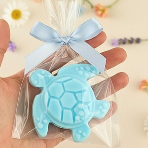 May include: A light blue sea turtle shaped soap bar packaged in a clear plastic bag. The bag is sealed at the top with a light blue satin ribbon tied into a bow. The soap features a detailed shell pattern and textured flippers on the surface.