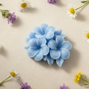 May include: A light blue, multi-petaled flower-shaped object with a smooth, molded texture. The object is surrounded by scattered small white daisies with yellow centers and small purple wildflowers on a textured, off-white fabric surface.