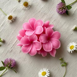 May include: A cluster of bright pink, molded wax flowers with smooth, rounded petals arranged in a dense, circular formation. The flower sculpture is surrounded by scattered white daisy-like flowers and small purple clover blossoms on a textured fabric.