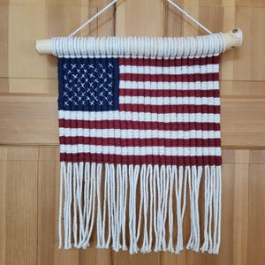 Puede incluir: Un tapiz de macramé que representa la bandera estadounidense. La bandera presenta un cuadrado azul con cruces blancas, rayas rojas y blancas y una franja. Está suspendido de un pasador de madera con una cuerda blanca.