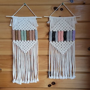 May include: Two macrame wall hangings with a diamond pattern and fringe. The left wall hanging has a brown, green, and pink color scheme. The right wall hanging has a black, pink, and gray color scheme.