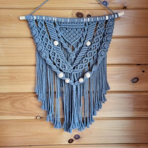May include: Gray macrame wall hanging with wooden beads. The macrame is in a geometric pattern with fringe on the bottom.