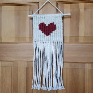 May include: White macrame wall hanging with a red heart woven into the center. The hanging has long white fringe.