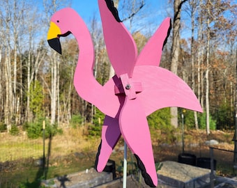 Puede incluir: Decoración de jardín de flamenco de madera rosa con pico amarillo y detalles negros. El flamenco tiene un diseño de molinillo con cuatro aspas rosas, montado sobre un poste de metal blanco. El fondo muestra árboles y cielo azul.
