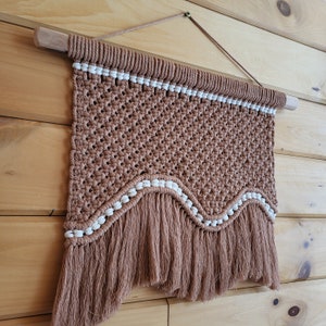 May include: A brown and white macrame wall hanging with a fringe border. The wall hanging is made with a knotting technique and has a wavy pattern.