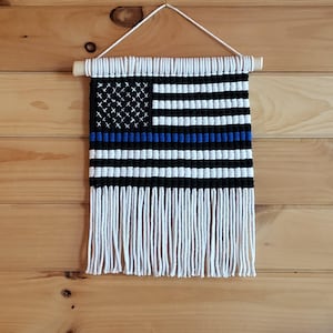 May include: A woven wall hanging in the shape of an American flag with a blue line running horizontally across the middle. The flag is made with white and black yarn and has a fringe at the bottom.