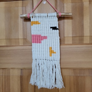 May include: White macrame wall hanging with pink, yellow, and black geometric shapes. The hanging has a fringe at the bottom.