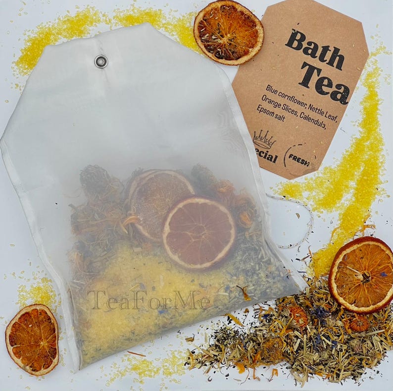 May include: A transparent bath tea bag filled with dried herbs and orange slices, with a brown tag that reads "Bath Tea". The tag lists ingredients like blue cornflower and Epsom salt. Dried orange slices and yellow granules are scattered around the bag.
