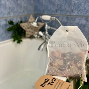 May include: A transparent bath tea bag filled with dried herbs and flowers, with the text "Tea For Me" printed on the bag. A brown tag with the words "Bath Tea" and ingredient details hangs from the bag. The background includes a bathtub, faucet, and a star-shaped decoration.