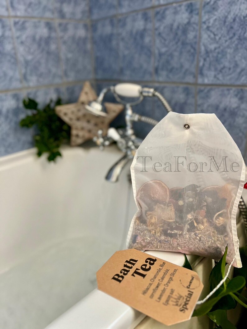 May include: A transparent bath tea bag filled with dried herbs and flowers, with the text "Tea For Me" printed on the bag. A brown tag with the words "Bath Tea" and ingredient details hangs from the bag. The background includes a bathtub, faucet, and a star-shaped decoration.