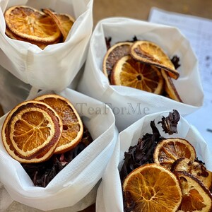 May include: Four white bags containing dried orange slices and dark red hibiscus flowers. The orange slices are a vibrant orange with a darker brown edge. The bags are on a wooden surface. The text "Tea for Me" is visible.
