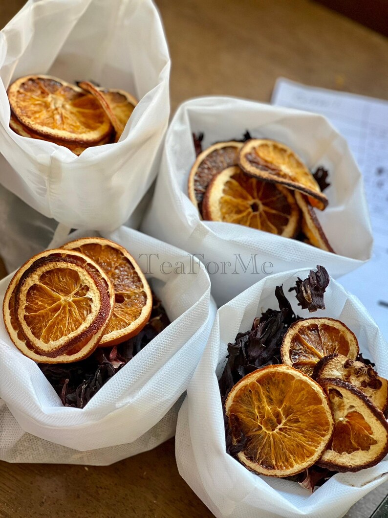 May include: Four white bags containing dried orange slices and dark red hibiscus flowers. The orange slices are a vibrant orange with a darker brown edge. The bags are on a wooden surface. The text "Tea for Me" is visible.