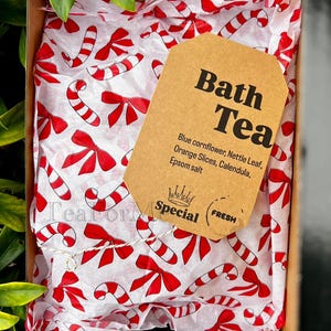 May include: A gift box containing bath tea. The box is lined with white tissue paper printed with red candy canes and bows. A brown tag reads "Bath Tea" with ingredients listed below. The box is surrounded by green foliage.