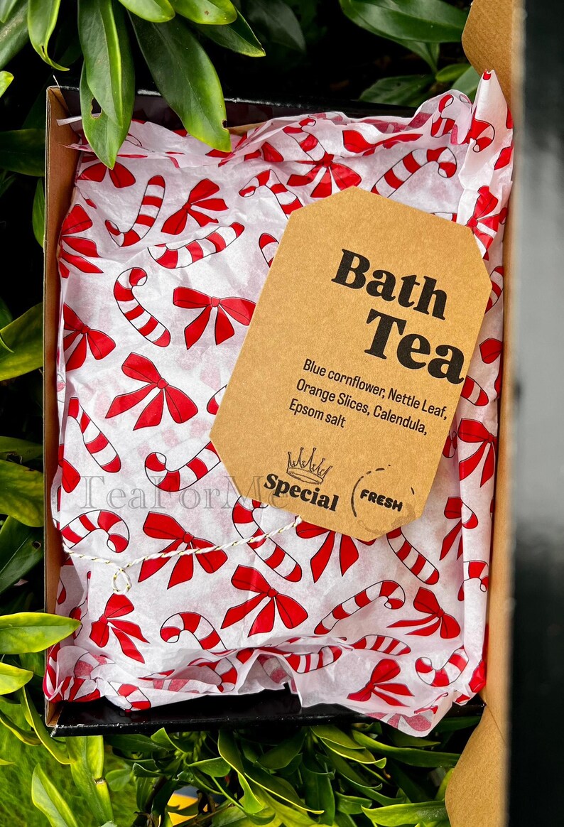 May include: A gift box containing bath tea. The box is lined with white tissue paper printed with red candy canes and bows. A brown tag reads "Bath Tea" with ingredients listed below. The box is surrounded by green foliage.