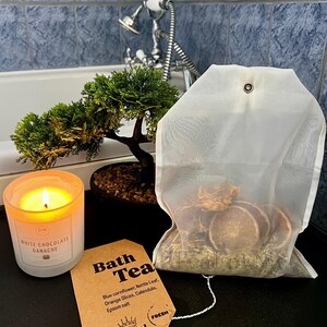 May include: A lit candle in a glass jar next to a bath tea bag and a small bonsai tree. The candle is labeled "WHITE CHOCOLATE GANACHE". The bath tea bag contains dried orange slices and herbs. A tag reads "Bath Tea".