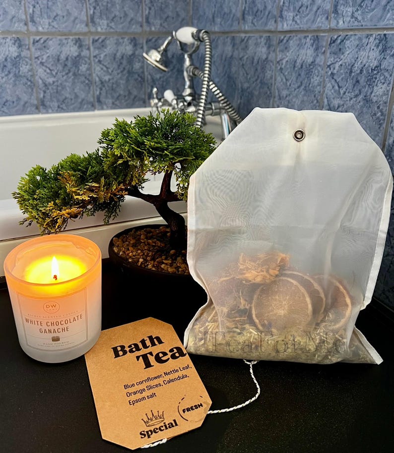 May include: A lit candle in a glass jar next to a bath tea bag and a small bonsai tree. The candle is labeled "WHITE CHOCOLATE GANACHE". The bath tea bag contains dried orange slices and herbs. A tag reads "Bath Tea".