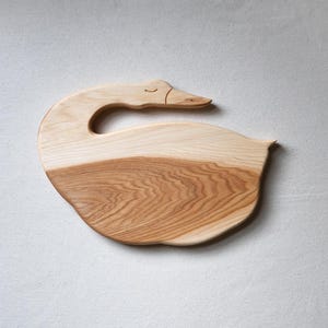 May include: A wooden cutting board shaped like a swan with a carved head and a handle. The board is made of light wood with a natural grain pattern.