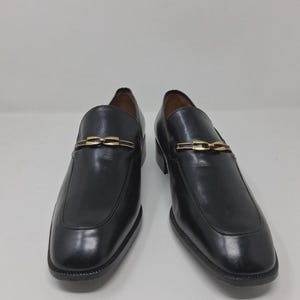 May include: A pair of black leather loafers with a gold-tone chain detail across the vamp. The shoes have a square toe and a low heel. The background is plain white.