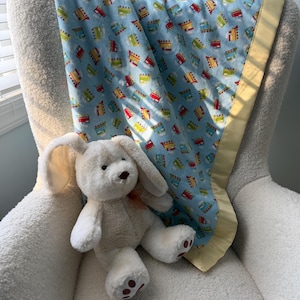 May include: A light blue baby blanket with a repeating pattern of colorful trains, trimmed with a yellow satin border. A white stuffed bunny with long ears and a pink bow sits on a white chair next to the blanket.