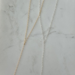 May include: Two delicate lariat necklaces, one in a gold tone and one in a silver tone, each featuring a fine cable chain design. The necklaces are constructed with a Y-shaped drop, where the chain splits into a single vertical strand.