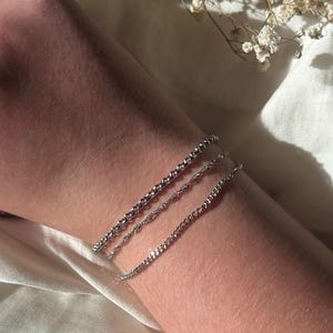 May include: Three silver bracelets of varying chain styles are displayed on a person's arm. The bracelets are delicate and shiny, reflecting light. The background is a soft, white fabric with small, dried flowers.