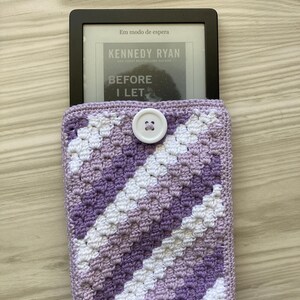May include: A purple and white crocheted e-reader case with a white button. The e-reader is inside the case and the screen displays the text "Em modo de espera" and the title of a book, "Before I Let."