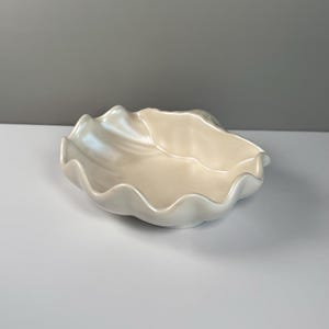 May include: A white ceramic bowl with a wavy, scalloped edge. The bowl is made of a smooth, glossy material and has a slightly iridescent finish.