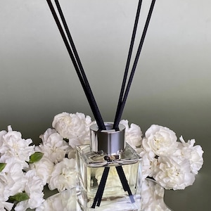 May include: A clear glass diffuser bottle with a silver cap and black diffuser sticks. The bottle is sitting on a silver, reflective, irregular-shaped tray. White carnations surround the bottle.