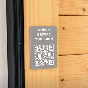 May include: A rectangular grey sign with rounded corners featuring the white text "CHECK BEFORE YOU BURN" above a white QR code. The sign is mounted on a vertical light-coloured wooden surface next to a black frame, providing clear safety instructions.