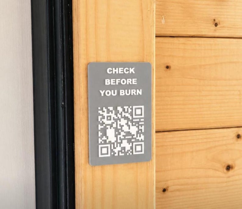 May include: A rectangular grey sign with rounded corners featuring the white text "CHECK BEFORE YOU BURN" above a white QR code. The sign is mounted on a vertical light-coloured wooden surface next to a black frame, providing clear safety instructions.