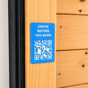 May include: A rectangular blue sign with rounded corners featuring the white text CHECK BEFORE YOU BURN and a white QR code. The sign is mounted on a vertical light-coloured wooden surface next to a black frame.