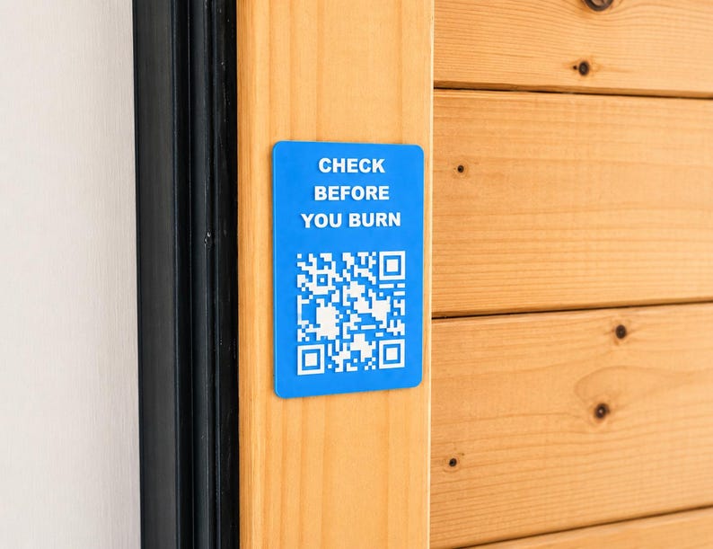 May include: A rectangular blue sign with rounded corners featuring the white text CHECK BEFORE YOU BURN and a white QR code. The sign is mounted on a vertical light-coloured wooden surface next to a black frame.