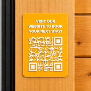 May include: A rectangular yellow sign with rounded corners featuring white raised text that reads VISIT OUR WEBSITE TO BOOK YOUR NEXT STAY! and a white raised QR code pattern centred below the text, mounted on a light wood surface.