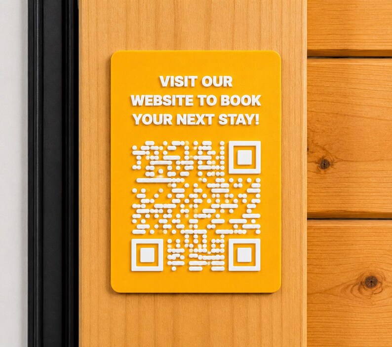May include: A rectangular yellow sign with rounded corners featuring white raised text that reads VISIT OUR WEBSITE TO BOOK YOUR NEXT STAY! and a white raised QR code pattern centred below the text, mounted on a light wood surface.