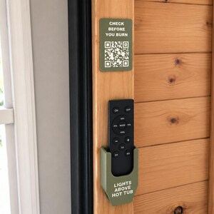 May include: A matte olive green plastic remote control holder mounted on a wooden frame. The holder features the white text "LIGHTS ABOVE HOT TUB" and contains a black remote. Above it, a matching sign reads "CHECK BEFORE YOU BURN" with a QR code.