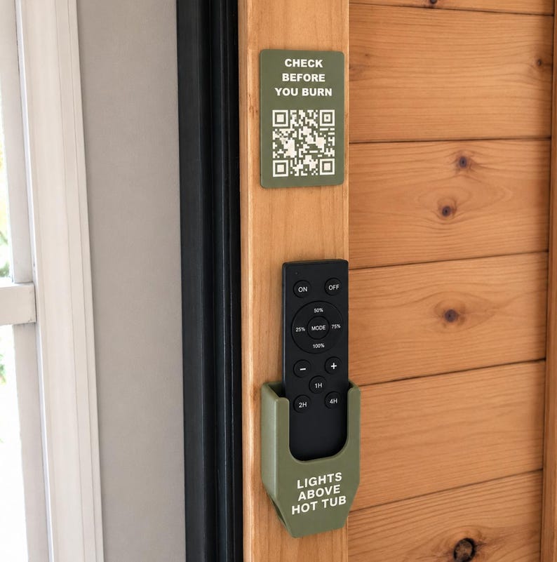 May include: A matte olive green plastic remote control holder mounted on a wooden frame. The holder features the white text "LIGHTS ABOVE HOT TUB" and contains a black remote. Above it, a matching sign reads "CHECK BEFORE YOU BURN" with a QR code.