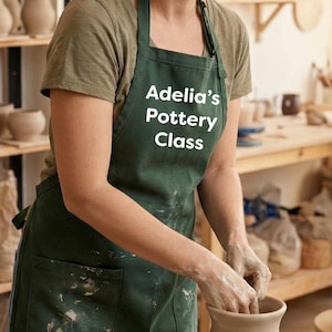 May include: A woman wearing a green apron with "Adelia's Pottery Class" printed on it, working on a pottery wheel. The pottery studio has shelves filled with finished pottery and bags of clay. The woman is smiling.