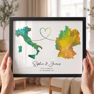 Può includere: Una stampa incorniciata con una mappa ad acquerello dell'Italia in verde e blu e della Germania in giallo e arancione, collegate da una linea sottile a forma di cuore. Il testo riporta Sophia & James, Italy & Germany, 28th September 2025.