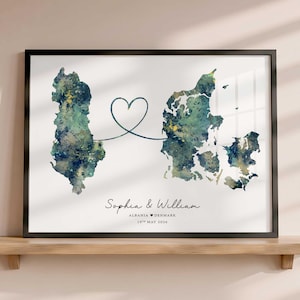 Op de afbeelding: Een ingelijste print met aquarelkaarten van Albanië en Denemarken verbonden door een donkerblauwe hartvormige lijn. Onder de kaarten staat de tekst Sophia & William, ALBANIA hart DENMARK en 19TH MAY 2026 in een strak, zwart lettertype.