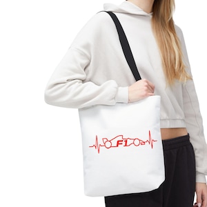 Puede incluir: Bolso tote blanco con una correa negra. El bolso presenta un gráfico rojo de un coche de carreras y el texto "F1" dentro de un diseño de línea de latido del corazón. El bolso es llevado.