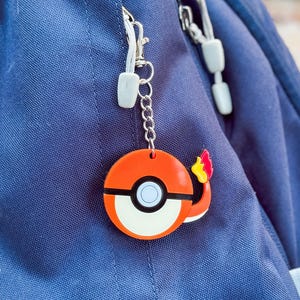 May include: A vibrant orange and white keychain featuring a cartoon design, attached to a blue backpack. The keychain has a circular shape with a black stripe and a small flame detail. The backpack has the Columbia logo.