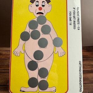 May include: A yellow and white game board with a cartoon illustration of a person with a red nose and a surprised expression. The board has eight gray circles on the person's body, labeled with ailments like "in stomach" and "broken heart". The text "AILMENT PARTS = $3" and "IF YOU GET BUZZED YOU OWE $5" is printed on the board. The text "CUTTING COST BUDGETING" is printed on the right side of the board.