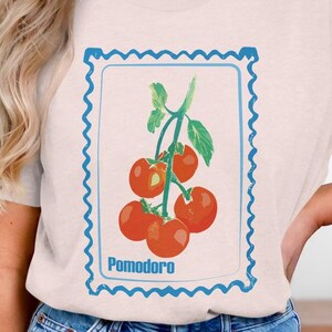 May include: A light beige t-shirt featuring a tomato illustration within a blue postage stamp frame. The illustration shows a cluster of red tomatoes with green stems. The word "Pomodoro" is printed below the tomatoes.