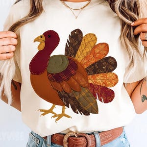 May include: Cream-colored t-shirt featuring a colorful turkey design. The turkey is made of various fabric patterns in shades of brown, orange, red, and yellow. The turkey has a red head and a yellow beak. The shirt is paired with blue jeans and a brown belt.