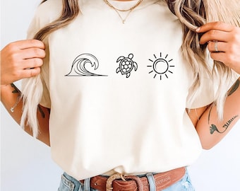 May include: Cream-colored t-shirt featuring three black line art illustrations: a wave, a turtle, and a sun. The shirt has a crew neck and short sleeves. The model is wearing blue jeans and a brown belt.