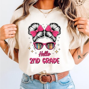 Puede incluir: Una camiseta color crema con un diseño gráfico para segundo grado. El diseño incluye la cara de una niña con lazos rosas, gafas de sol con lápices de colores arcoíris y las palabras "Hello 2nd Grade" en rosa y negro.