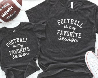 May include: Two gray t-shirts with the text "Football is my favorite season" on them. The smaller shirt is for a child and the larger shirt is for an adult. A brown and white football is in the background.