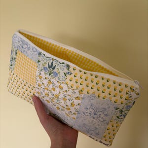 May include: A patchwork zippered pouch with a white zipper and a yellow gingham interior. The exterior features floral and patterned fabric in yellow, white, and blue. The words "Pixie & Thread" are printed on the side.